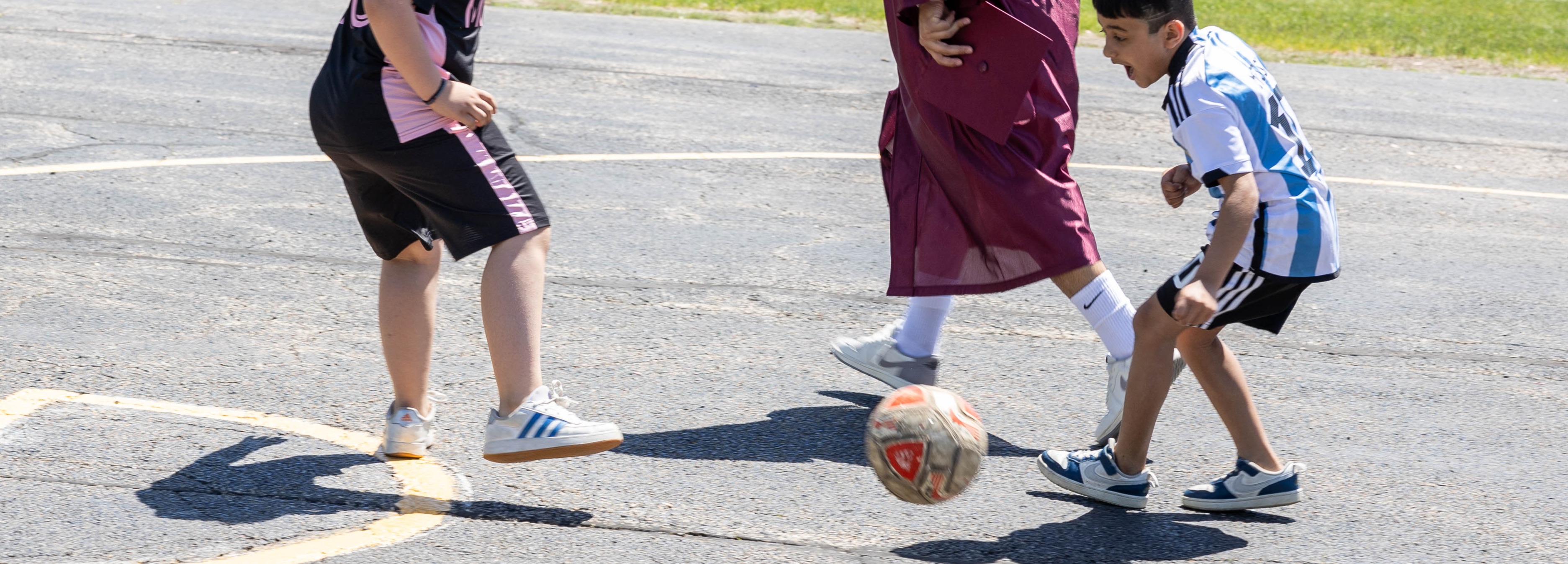 Three individuals on a paved area, two children playing with a ball, one adult observing, background includes grass, trees, and a sports field.
