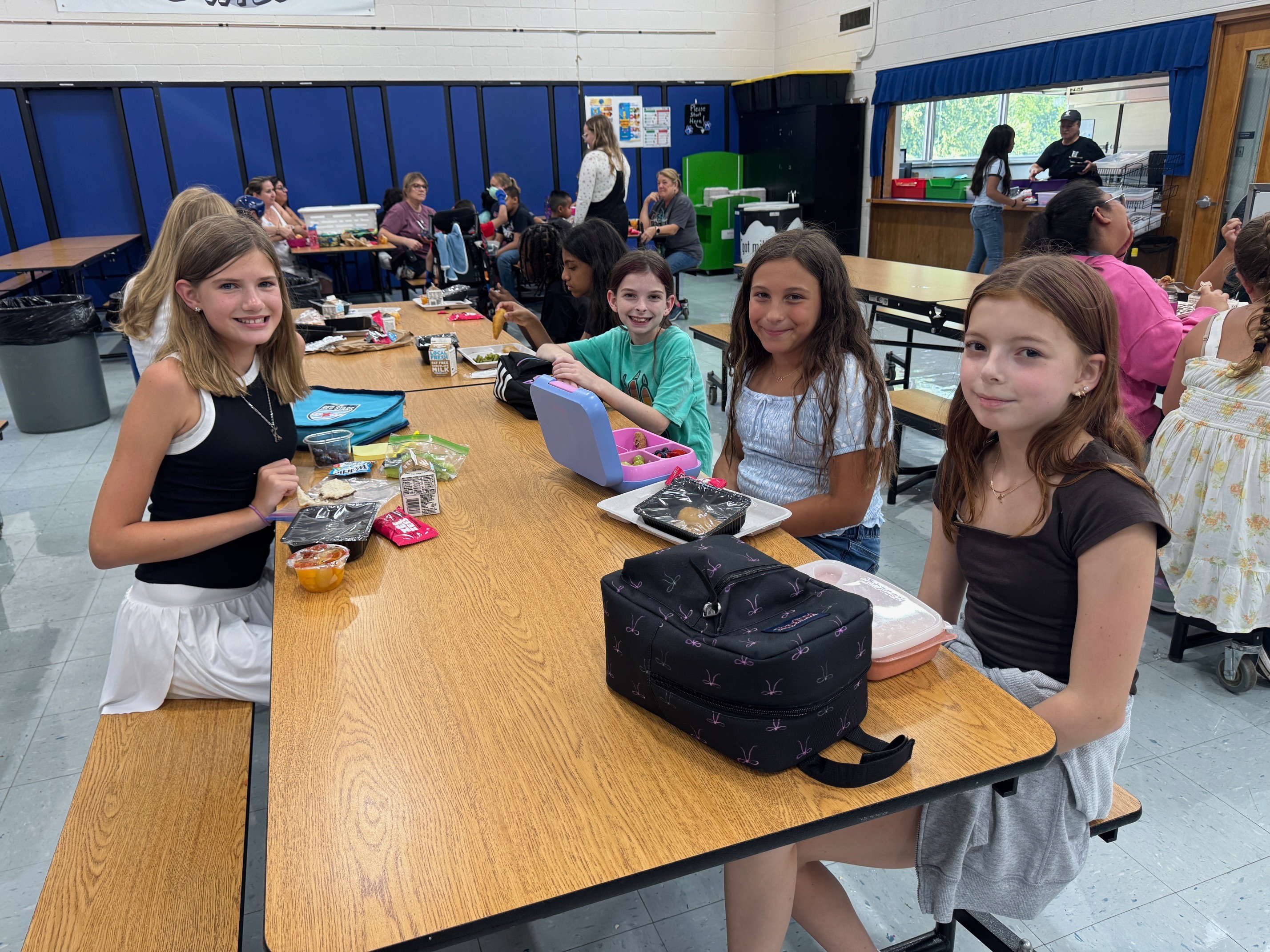 A group of girls sit at a table in a cafeteria. One has a black backpack on the table.