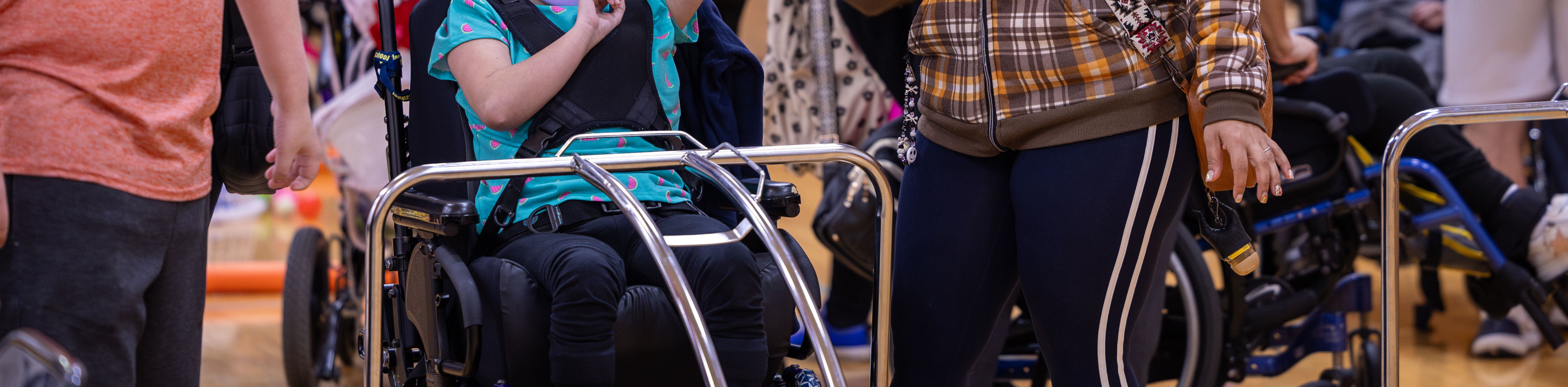 Several people in a gym, including a person in a wheelchair raising their hand, and others standing and watching.
