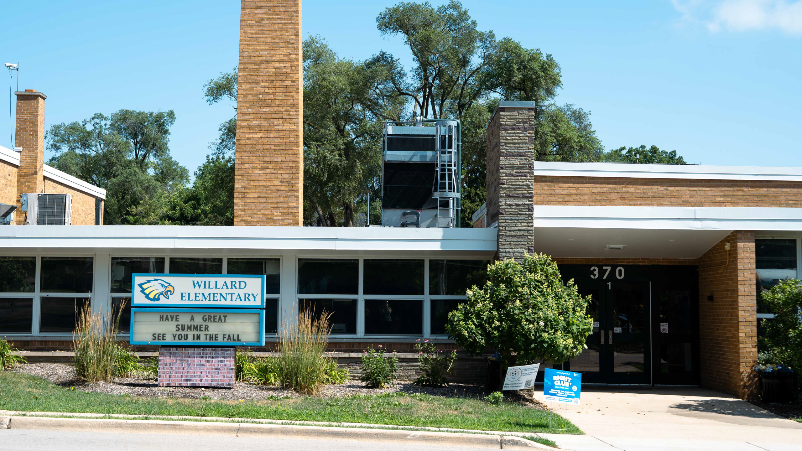 Exterior of a school building with a brick facade, tall chimneys, large windows, and a signboard.