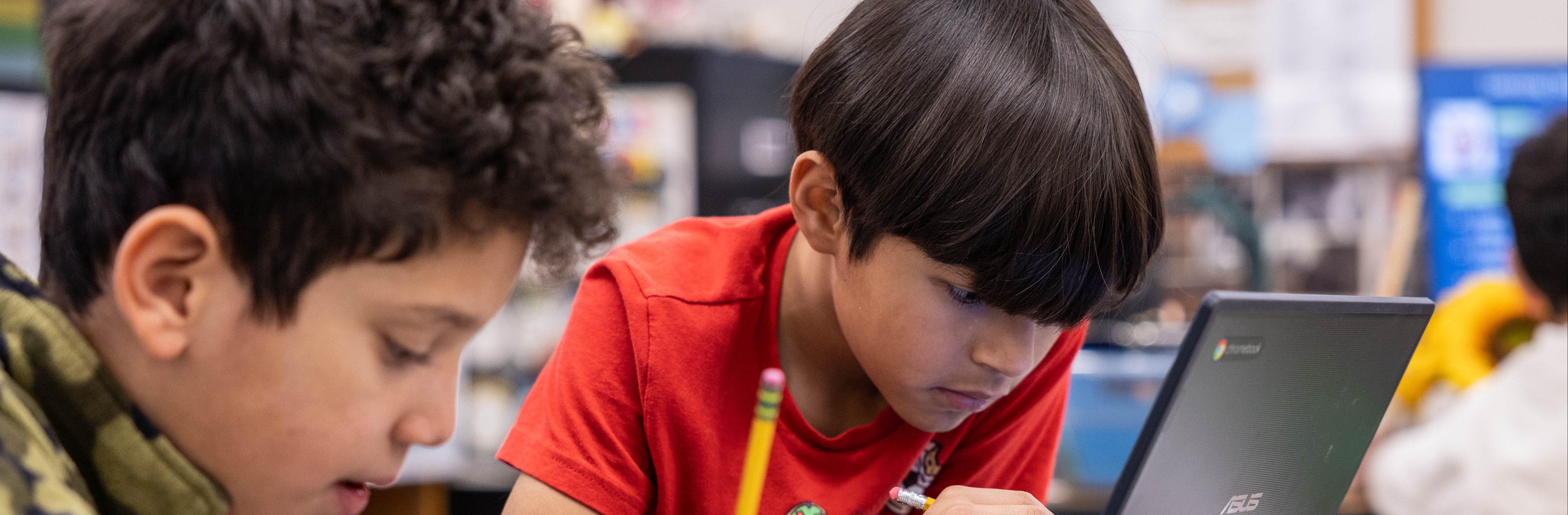 Two boys in a classroom, one in a camouflage jacket, the other in red, both focused on a laptop.