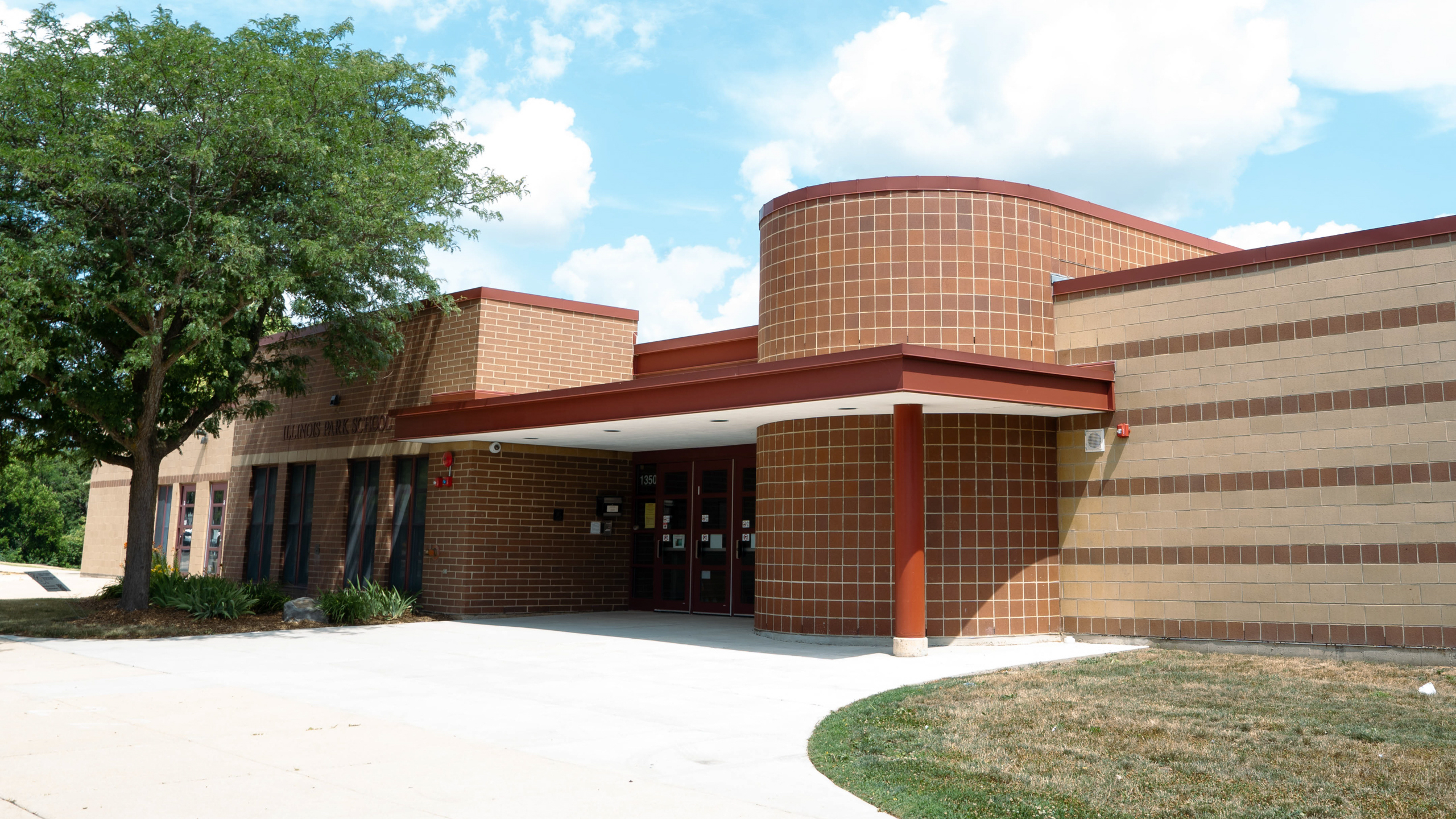 A brick building with a circular section in front. A tree stands on the left. A walkway leads to the entrance.
