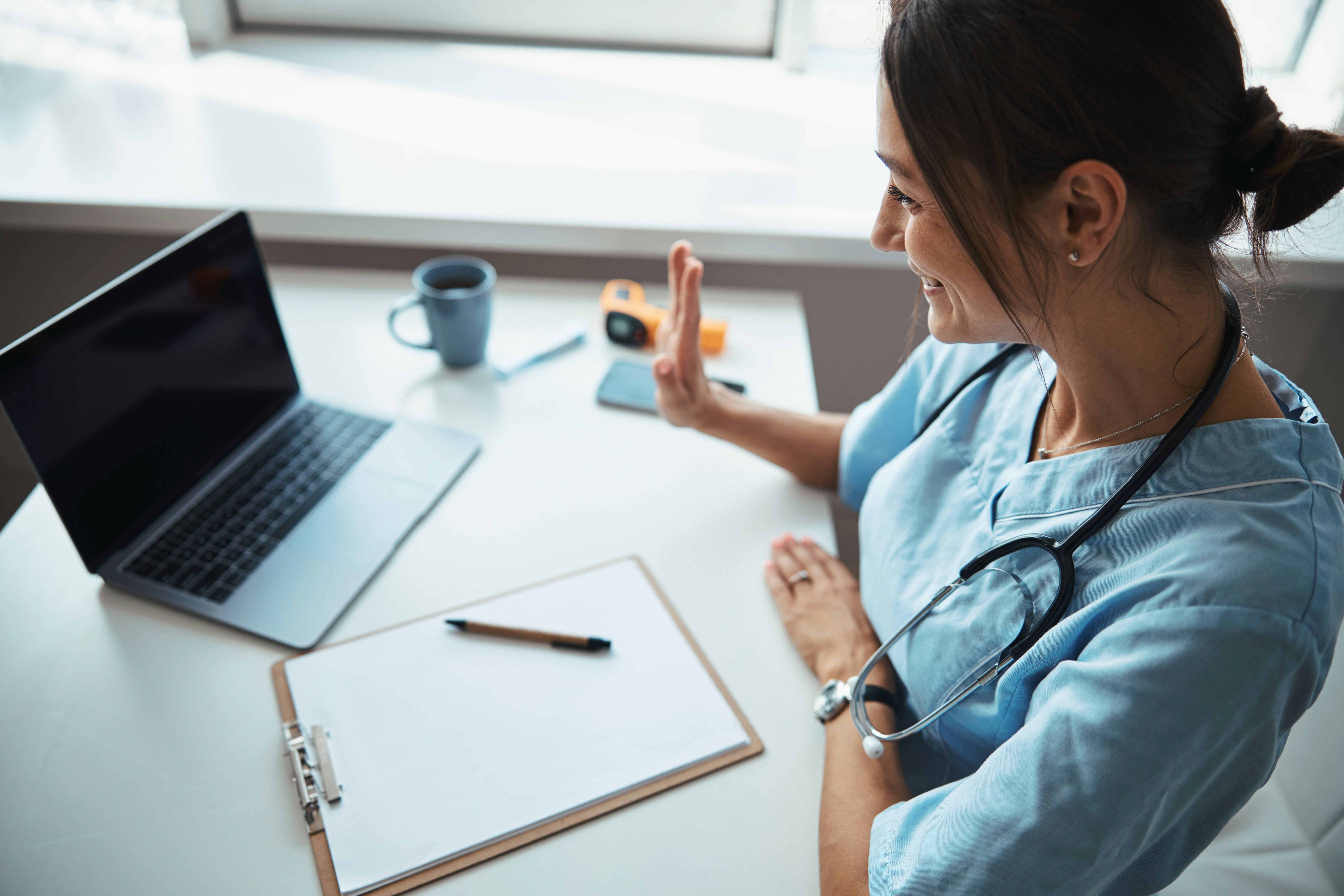 A woman in blue scrubs with a stethoscope around her neck, seated at a desk with a laptop, a pen, and a cup.
