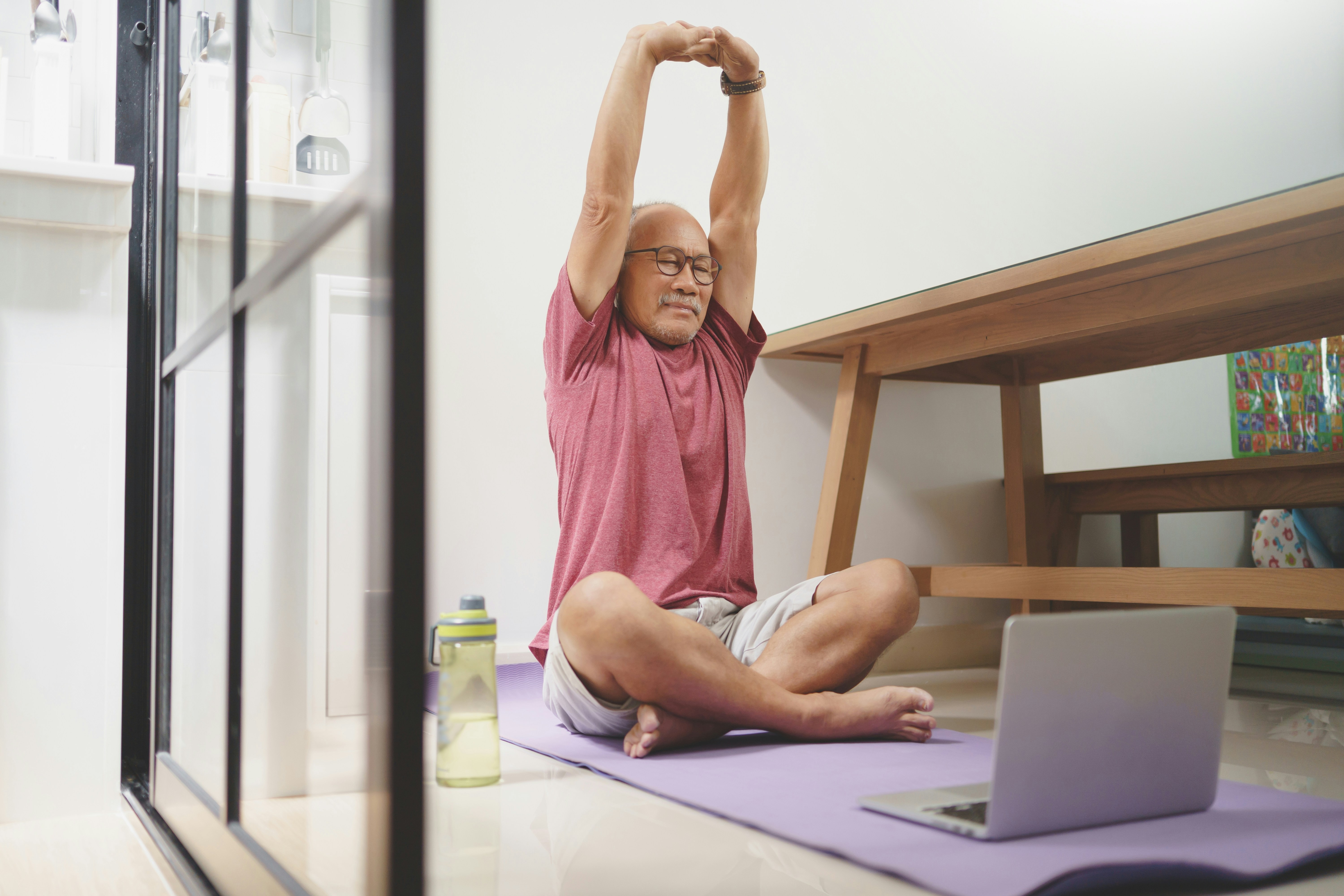 A man in glasses and a pink shirt doing yoga with a laptop and water bottle.