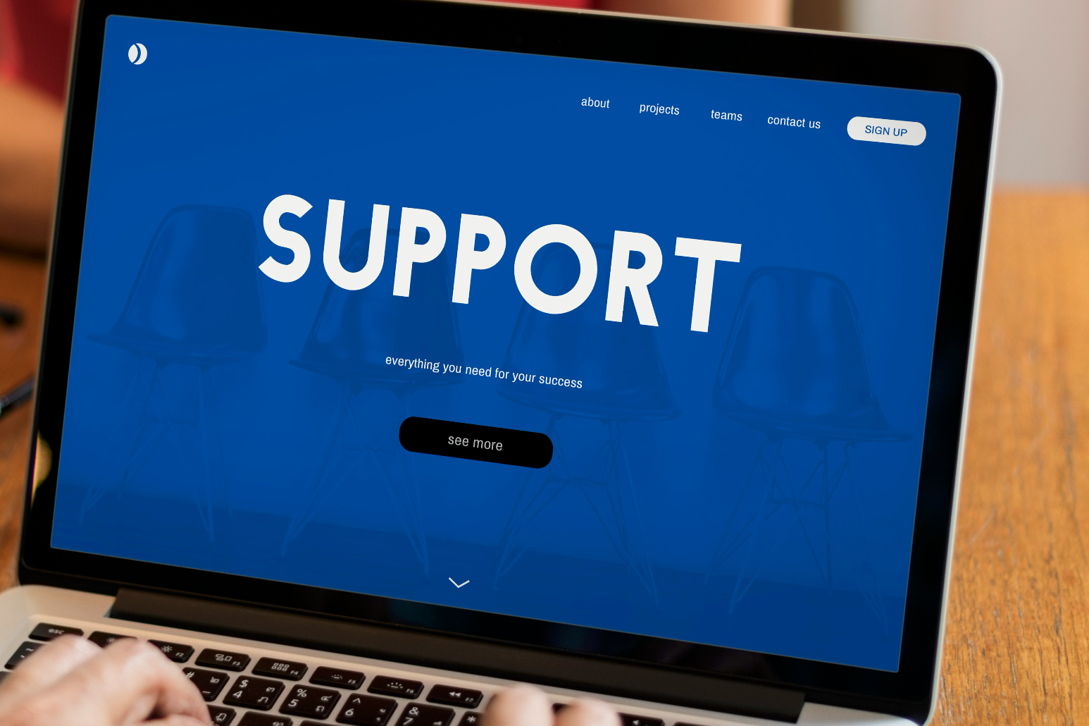 Person typing on a laptop with a blue screen displaying the word "SUPPORT" on a wooden desk.