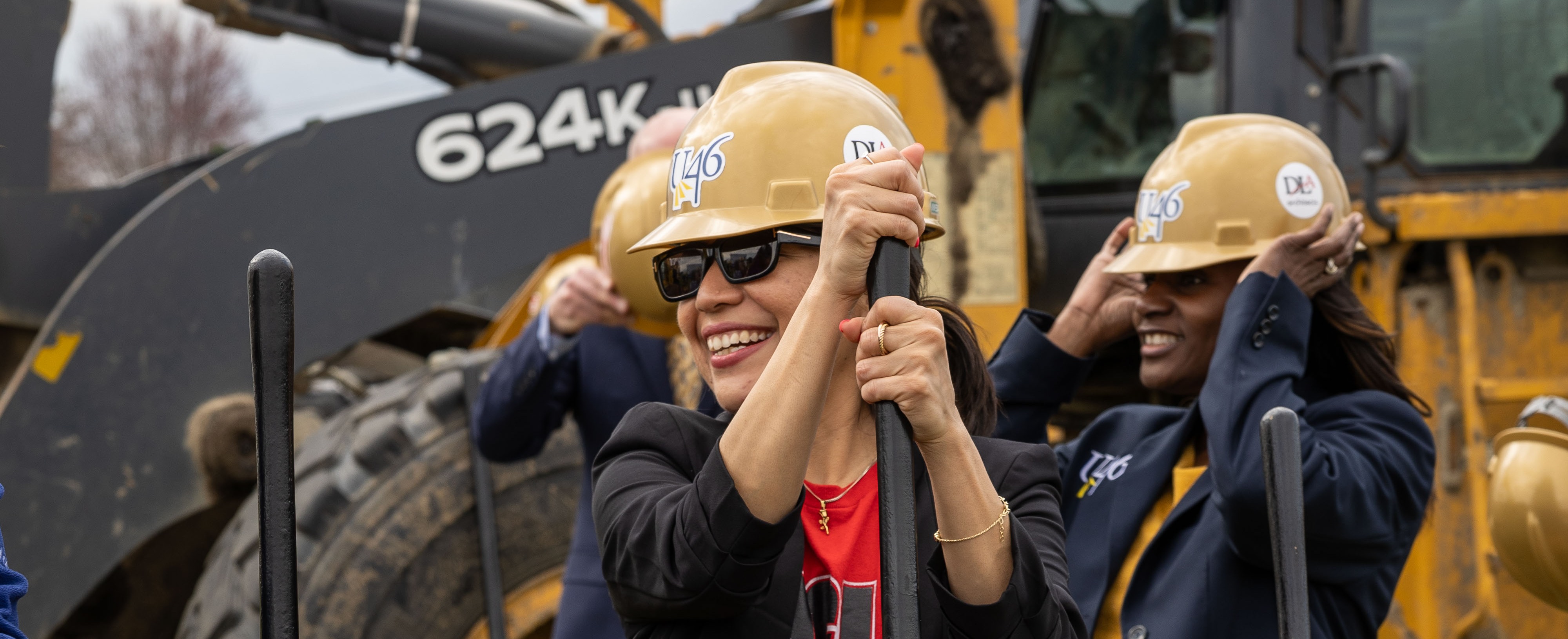 Three women wearing hard hats hold shovels at a construction site. Behind them, a large bulldozer rests on a grassy area.