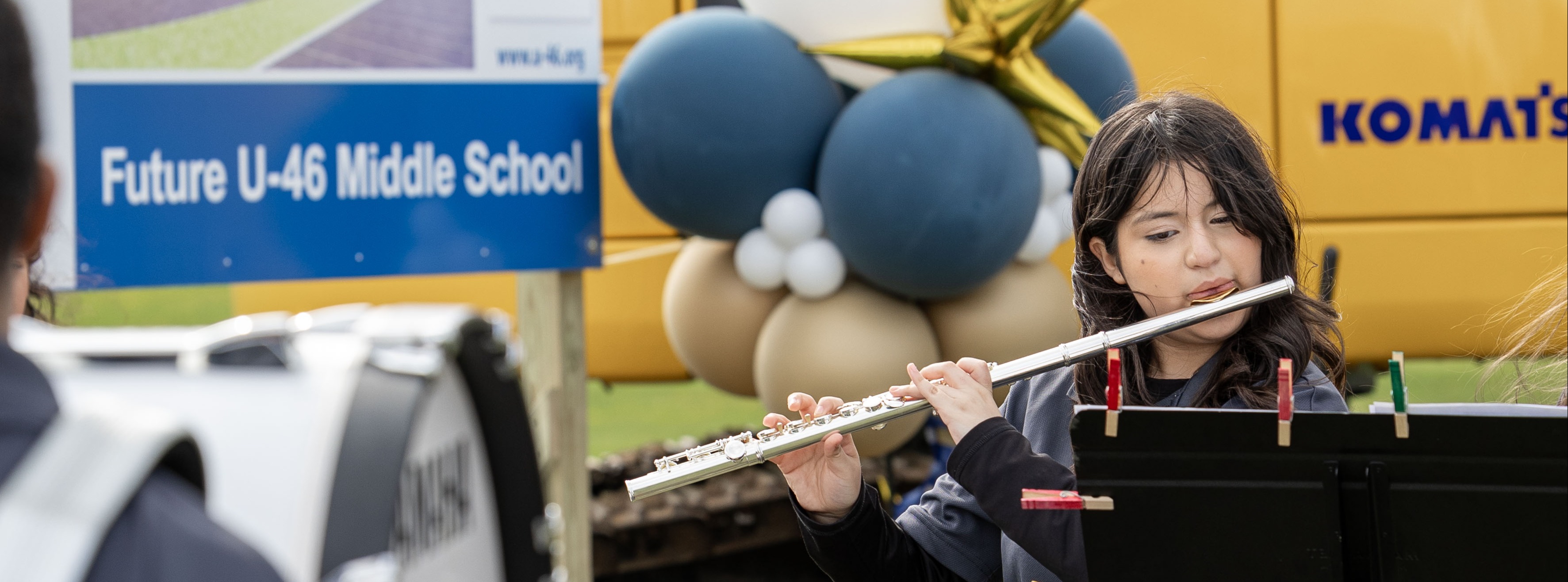 A young woman plays a flute while a man holds a drumstick, both performing in front of a yellow excavator.