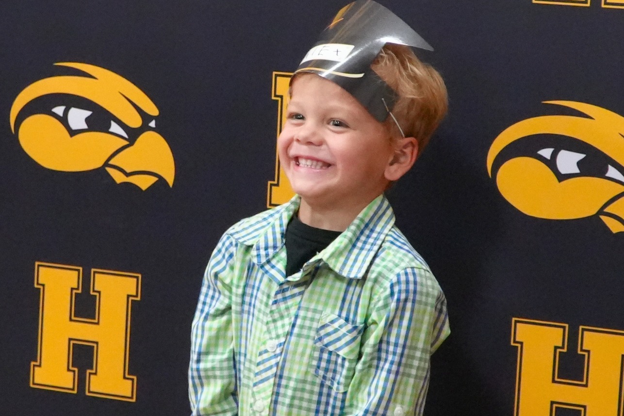 A young male preschool graduate smiles in front of a Hamilton backdrop.