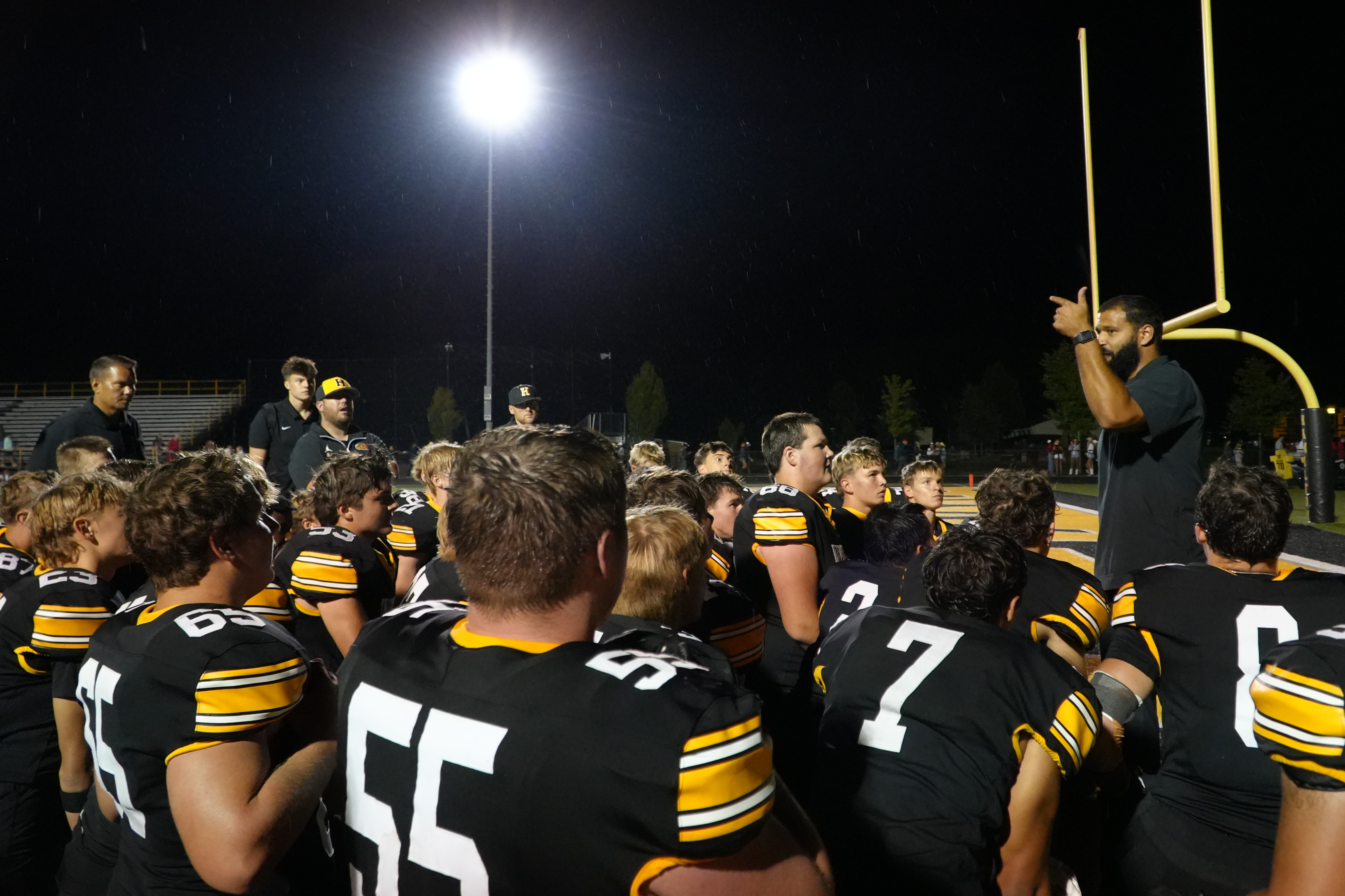 A coach addresses a football team in black and gold uniforms in darkness under a light.