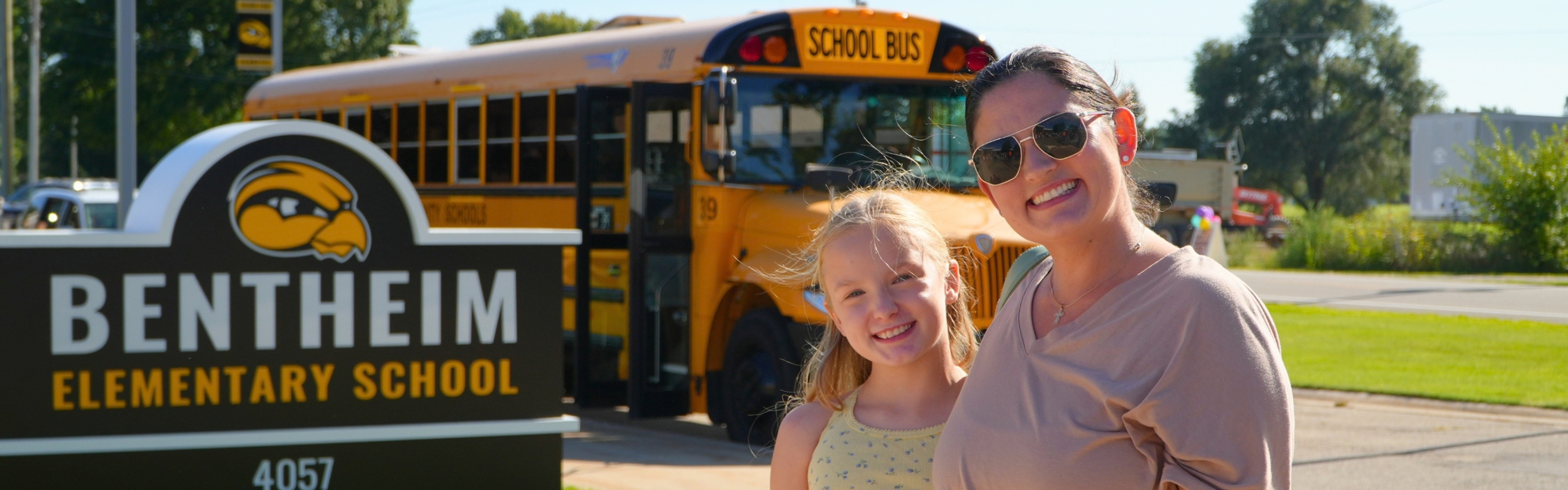 A female student with a female adult smile in front of a school bus and Bentheim Elementary sign.