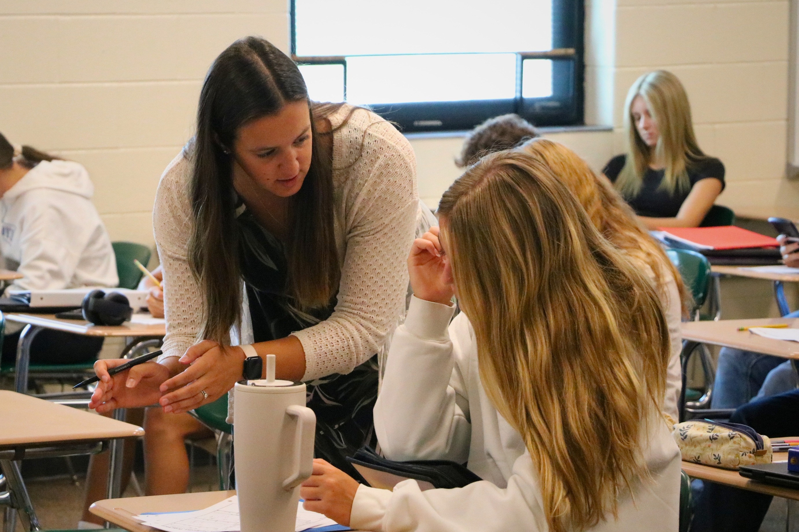 A female teacher helps a student sitting at her desk.