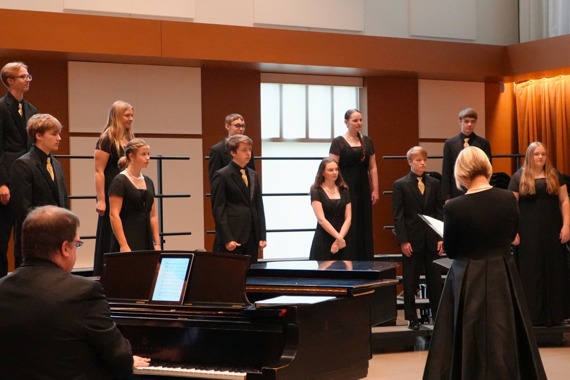 A group of students in all black prepares to sing with a piano and teachers.