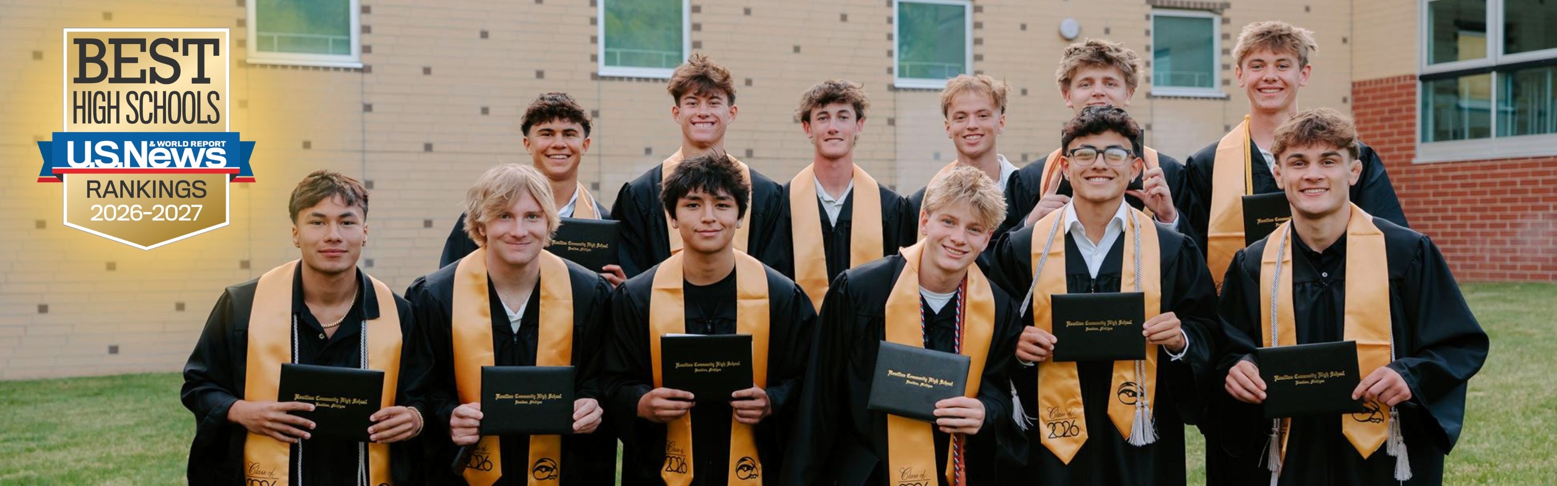 A group of male graduate students stand in front of a brick school in black and gold.