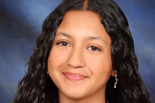Person with long, curly black hair wearing a black top, a necklace with a pendant, and earrings.