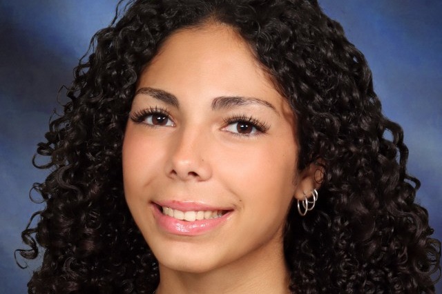 A person with curly dark hair wearing a black top, a necklace with a heart pendant.