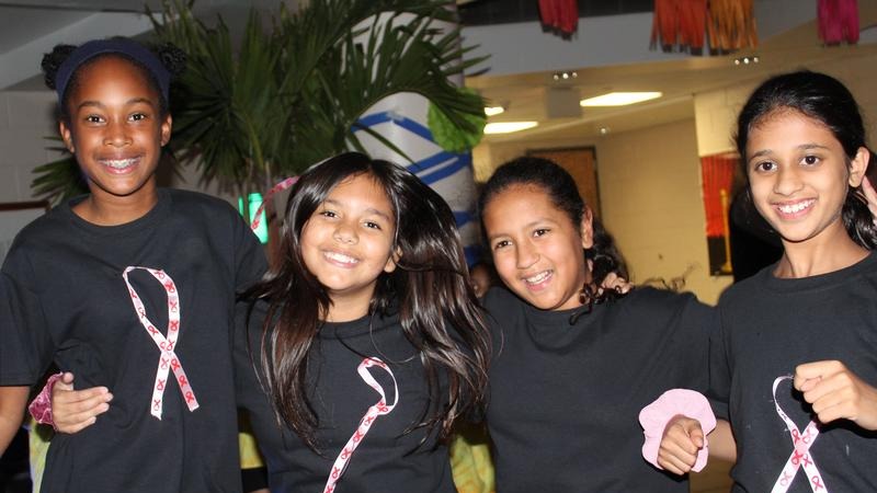 Four girls in black t-shirts with pink ribbons stand together in a room, smiling and posing.