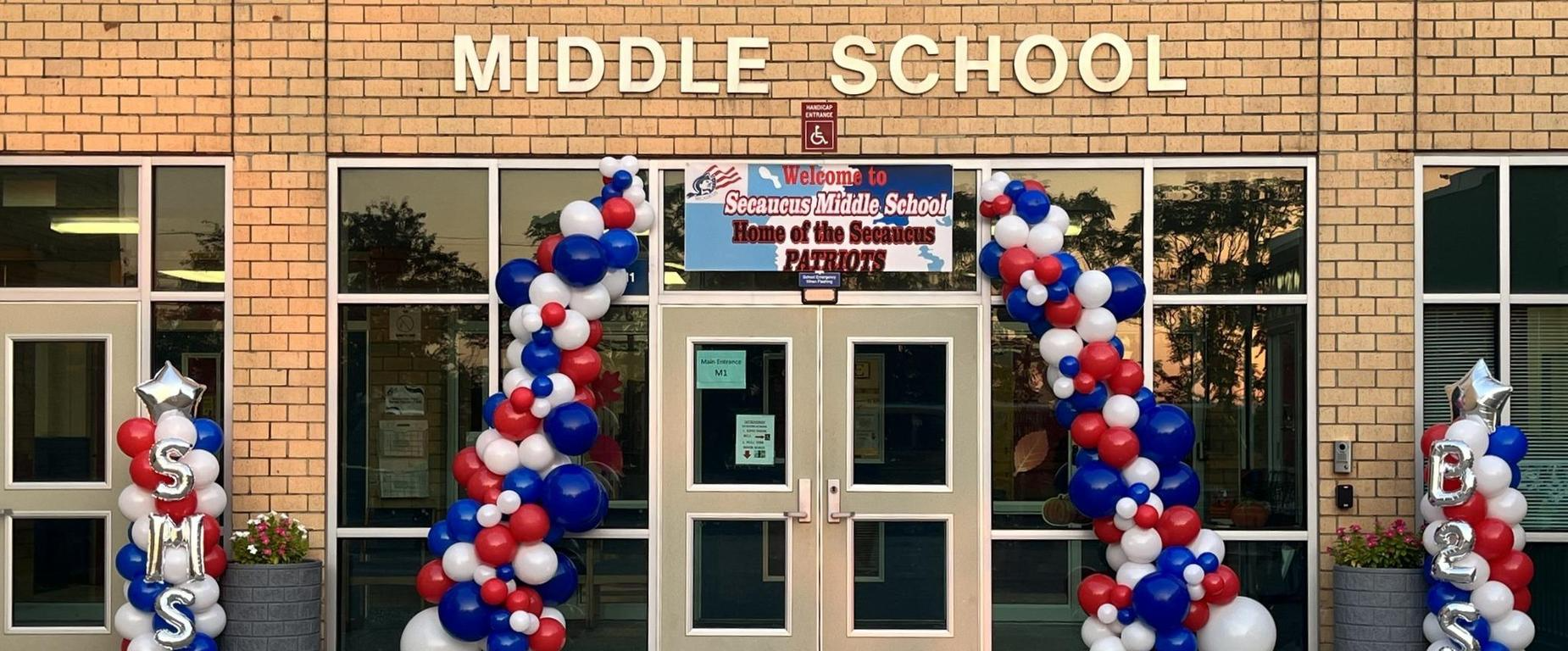 A middle school entrance adorned with red, white, and blue balloons. Glass doors and windows reflect trees.