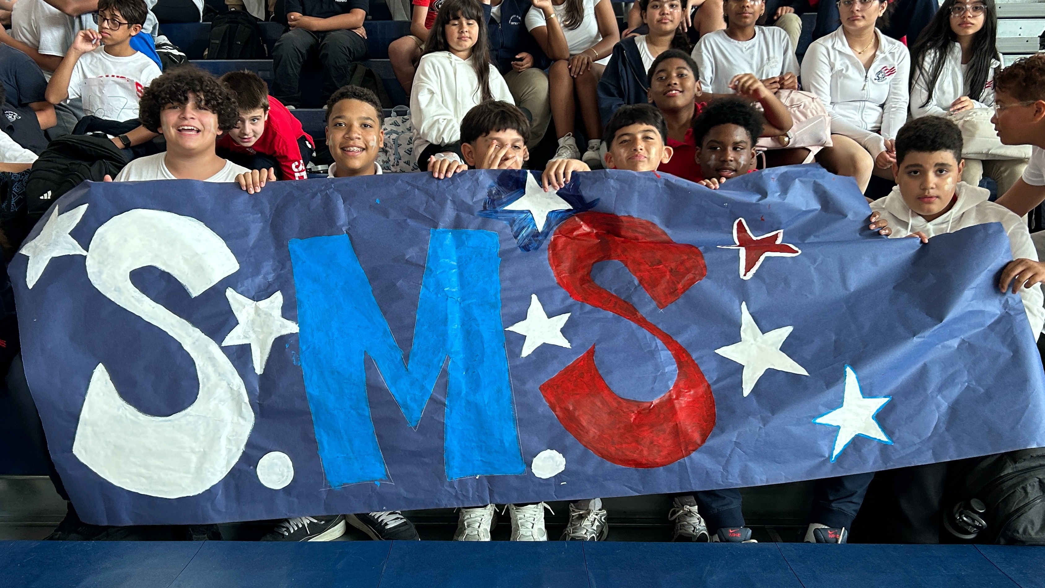 A group of children in school uniforms in bleachers, holding a banner reading SMS in front of them.