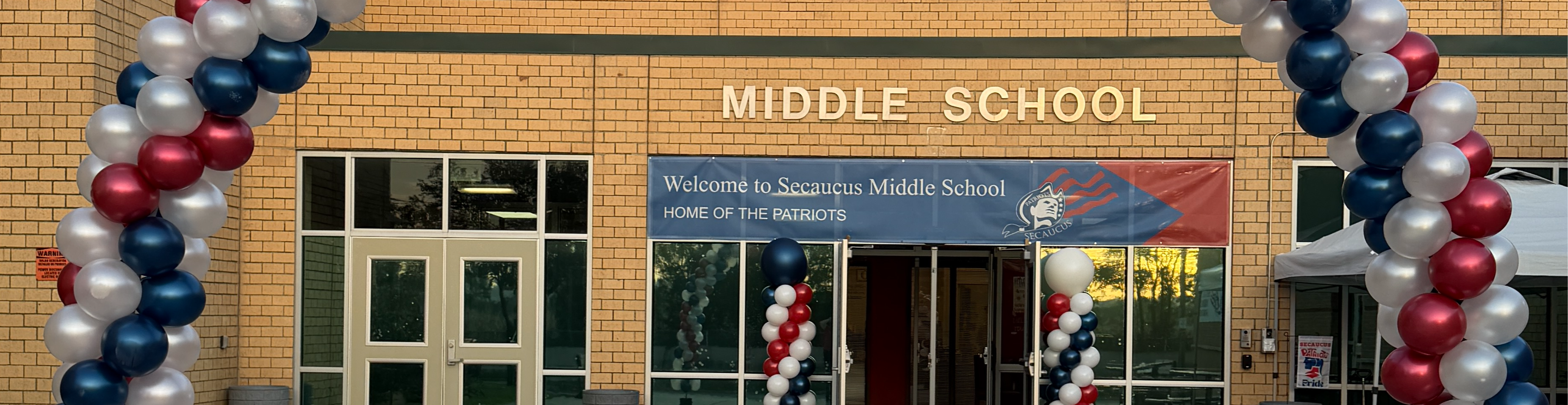 A middle school entrance adorned with red, white, and blue balloons. Glass doors and windows reflect trees.