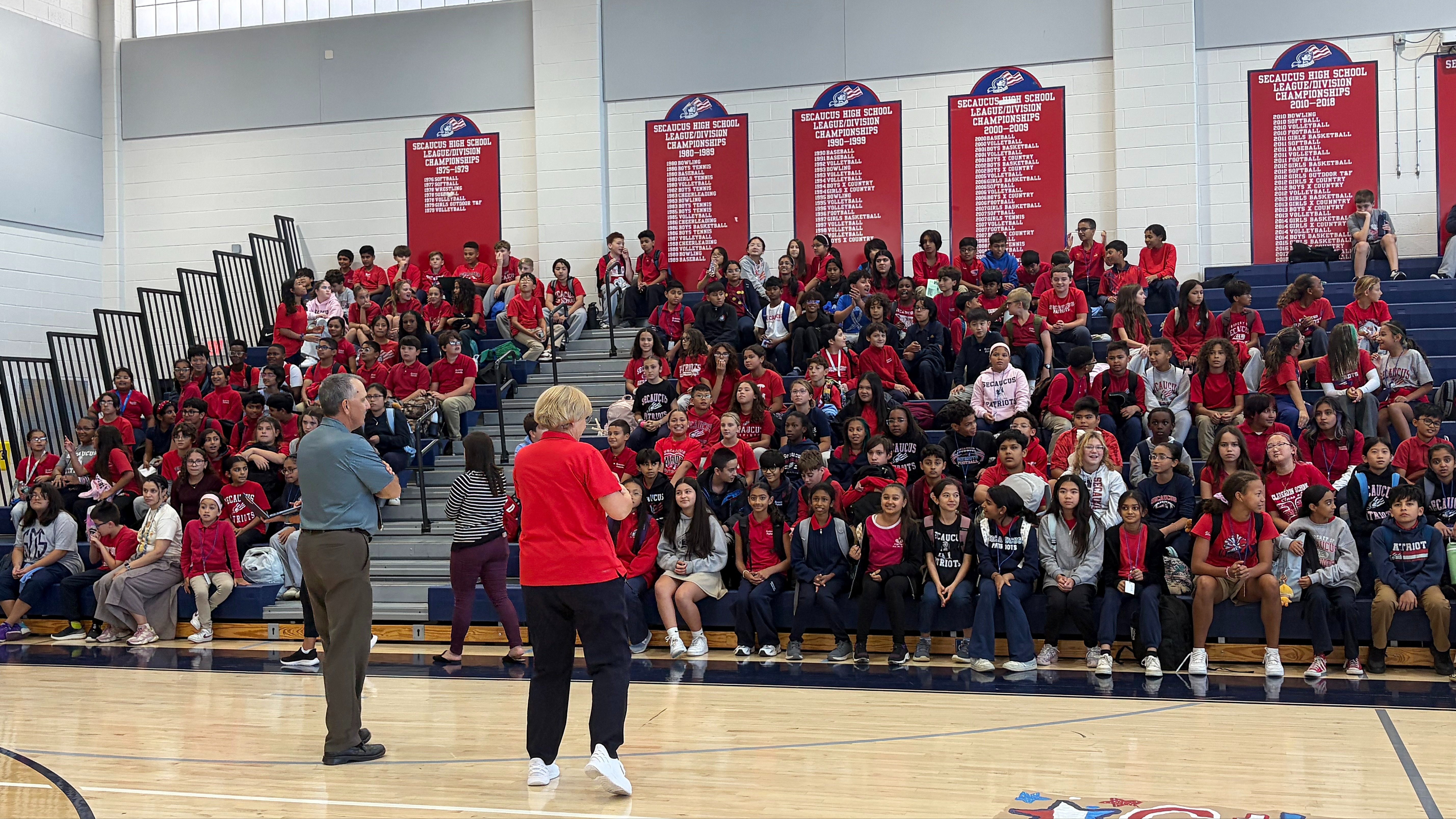 A large group of people gathered in a gym. Two people are standing at the front. Banners are on the wall.