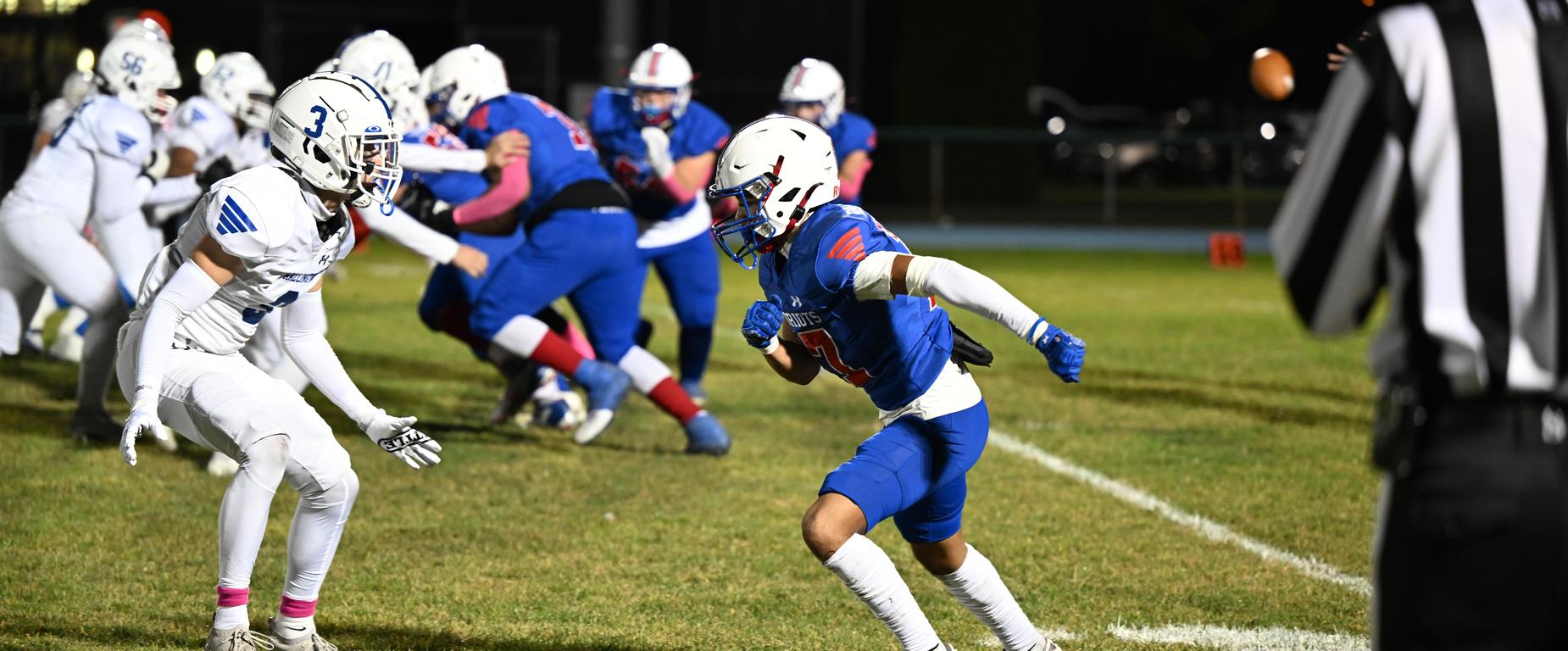 Several players in blue uniforms are running on a field, one carrying a football. An official stands nearby.