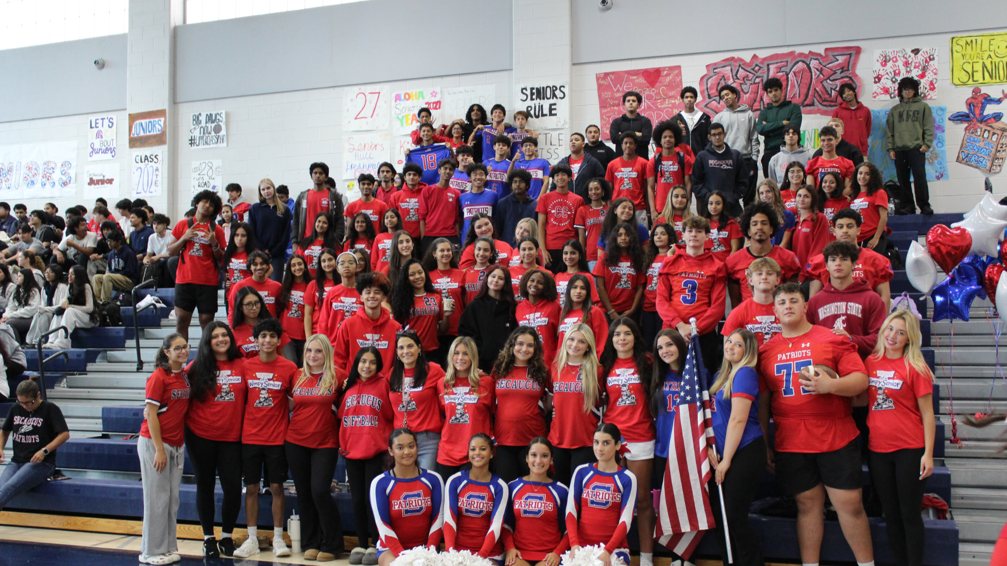 A large group of students in red and white shirts gather on gym bleachers, with some holding American flags, in a school gymnasium.