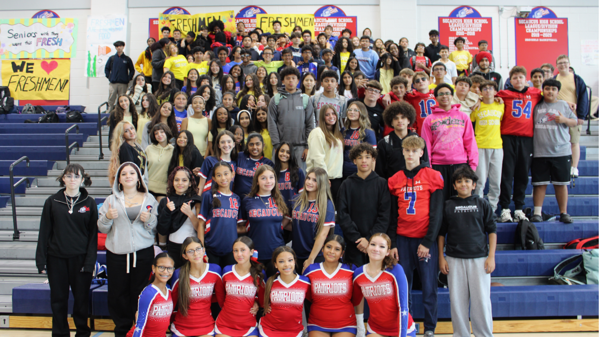 A group of cheerleaders, athletes, and adults pose for a group photo on gymnasium bleachers, with a colorful banner and spectators in the background.