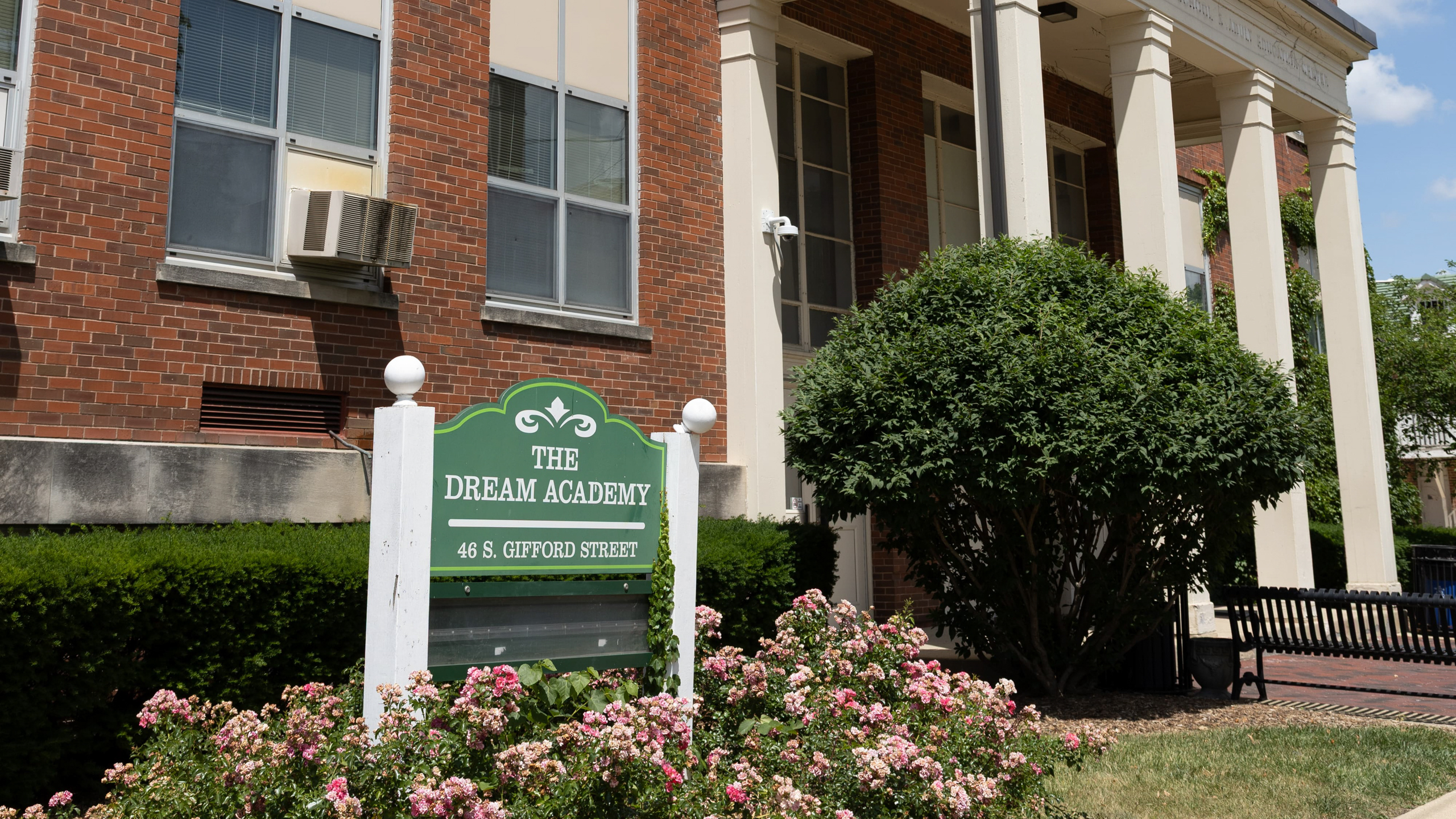 Brick building with columns, sign reading "The Dream Academy" and greenery in front.