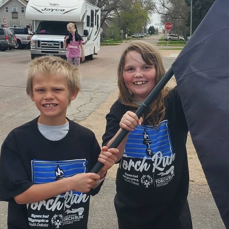 Two elementary students are carrying a flag and wearing black tee shirts with white lettering that says Torch Run