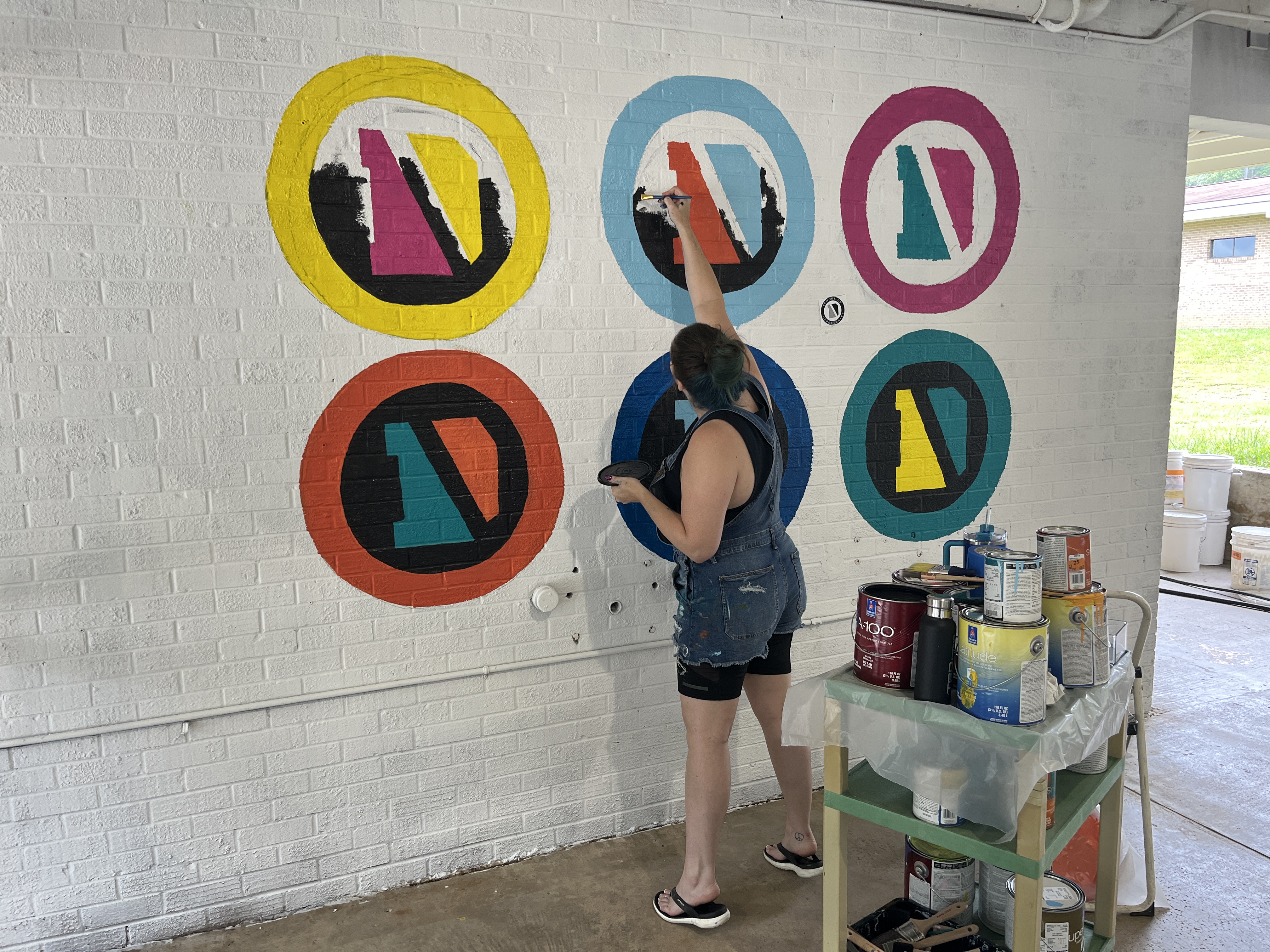 Woman painting mural of DRIVEN logos in many colors on school wall