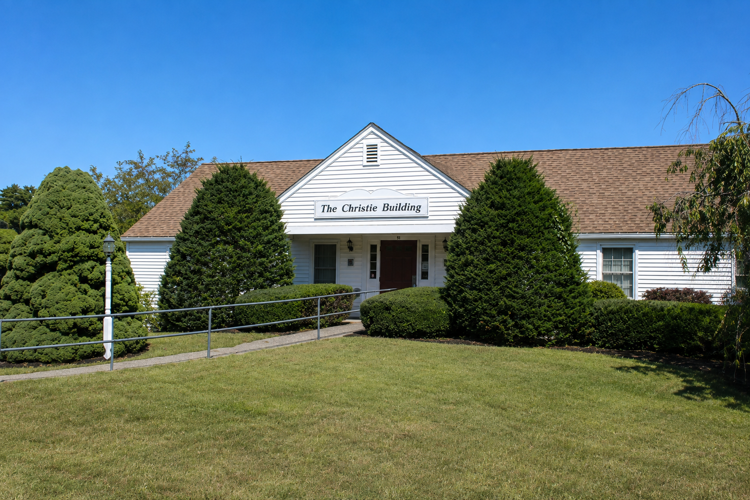 Exterior view of the SAU 17 Central Office in the Christie Building, a white, single-story building with a brown shingled roof and a central covered entrance. Evergreen trees and trimmed shrubs frame the entrance, with an accessible walkway and railing crossing the lawn beneath a clear blue sky.