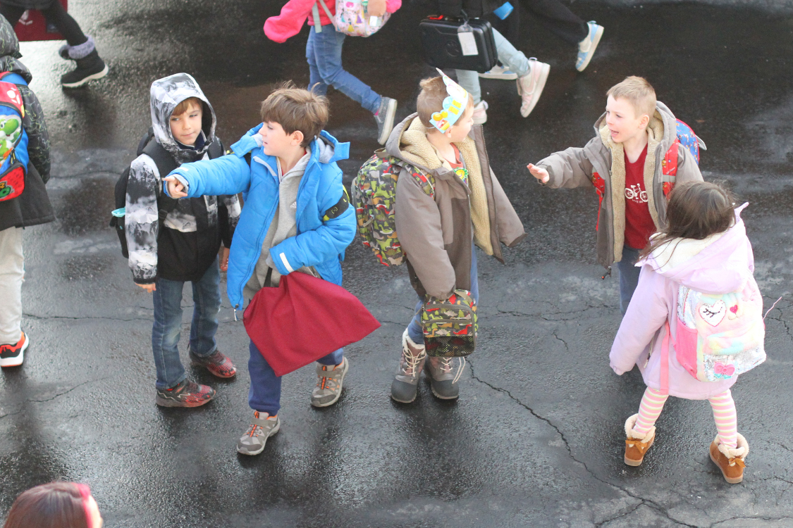 Multiple children in winter coats stand in a wet area, some carrying backpacks. Background shows wet pavement.