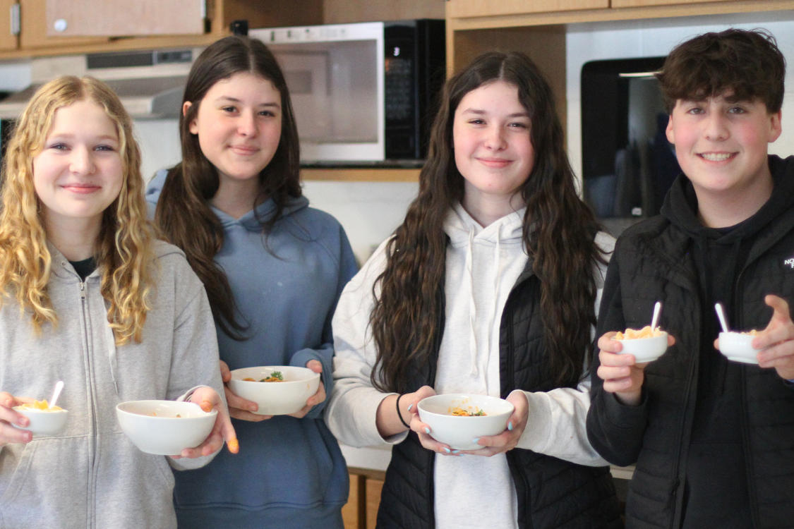 Five young people, three girls and two boys, smile and pose in a kitchen while holding bowls.