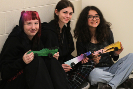 Three women sit against a white wall, each holding a toy guitar. One wears pink hair, another glasses, and the third has long hair.