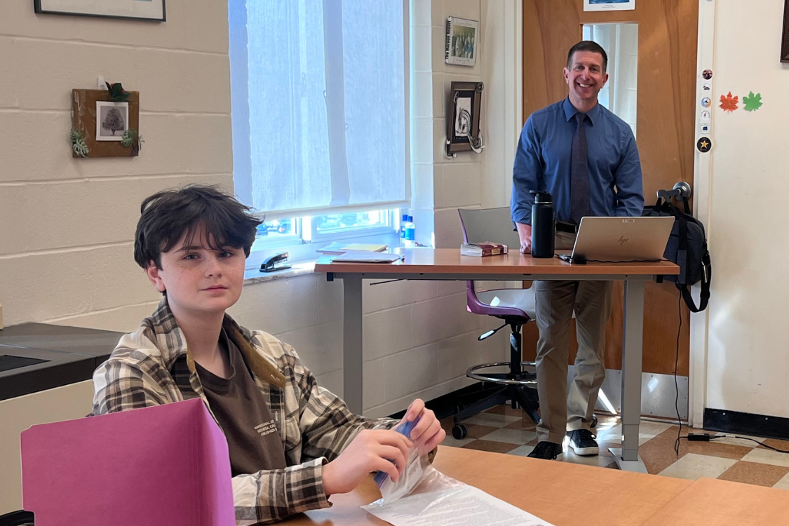 A student sits at a desk with papers and a tablet. A teacher stands at a desk behind him.