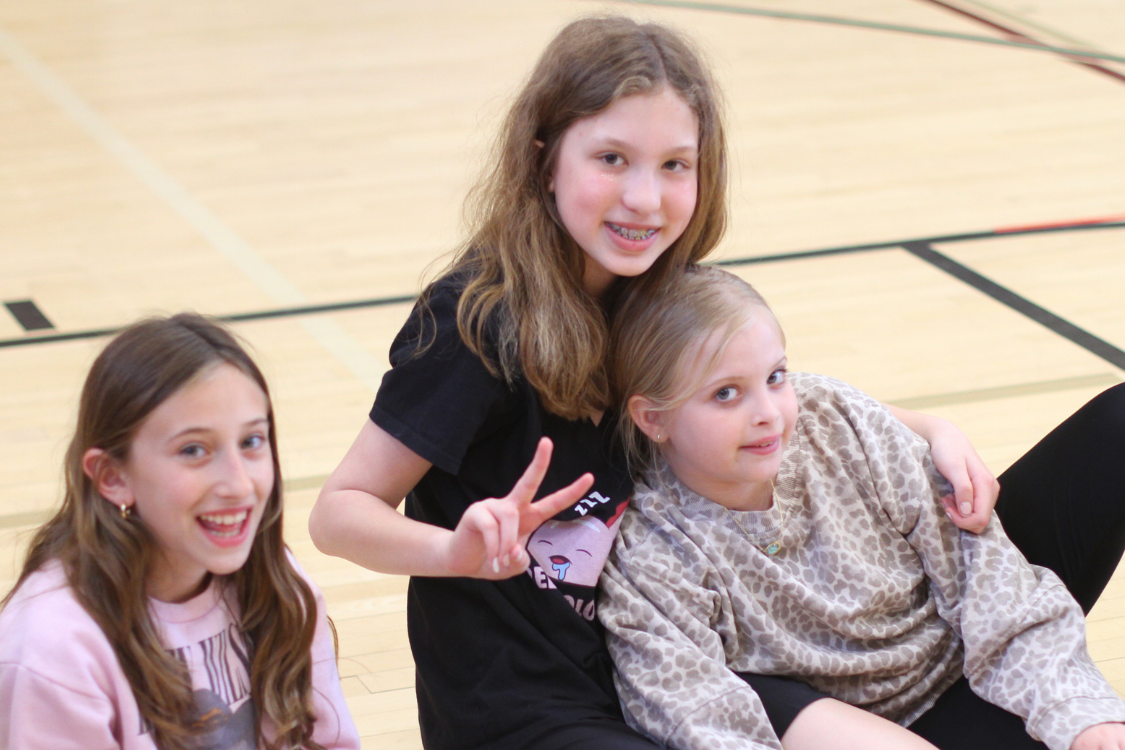 Three girls in a gymnasium, two sitting on the floor and one leaning on them, all smiling.