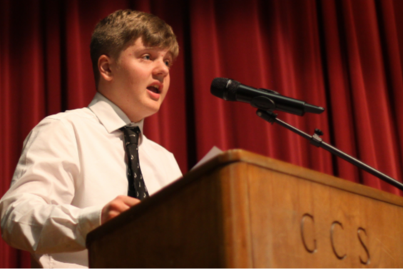 Person speaking at a podium with microphone; red curtain backdrop. Text on podium: CCS.