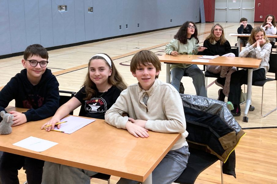 Several students are seated at desks in a gym, focusing on their papers. A wooden floor and gray walls are visible.