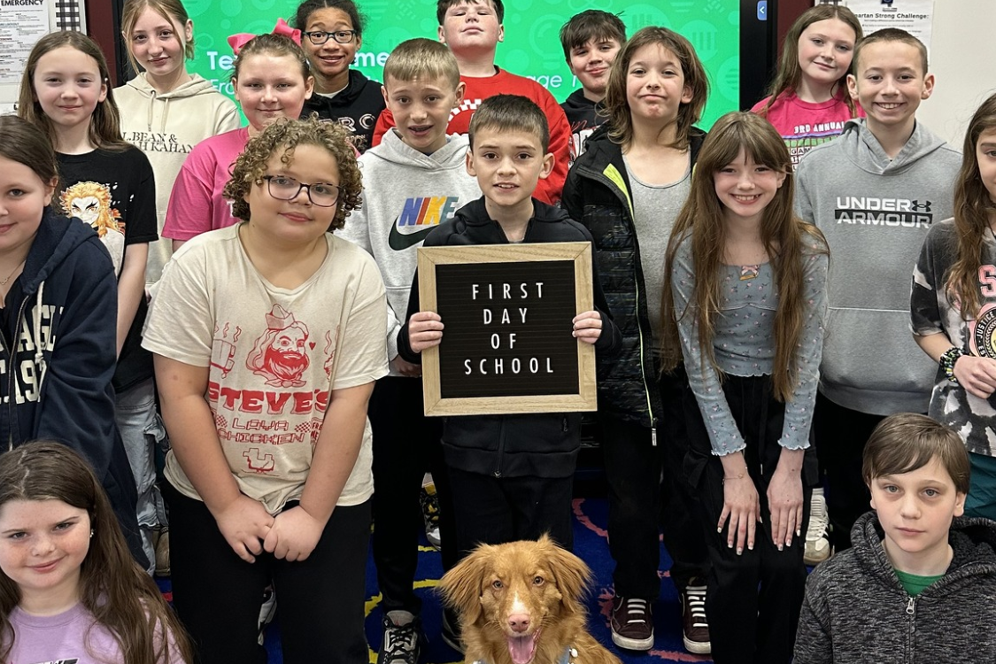 A classroom with children standing in two rows; a boy holds a framed certificate, and a dog stands nearby.