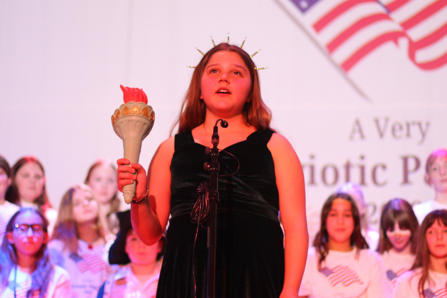A woman holding a torch and a crown stands on stage. Children in white shirts stand behind her.