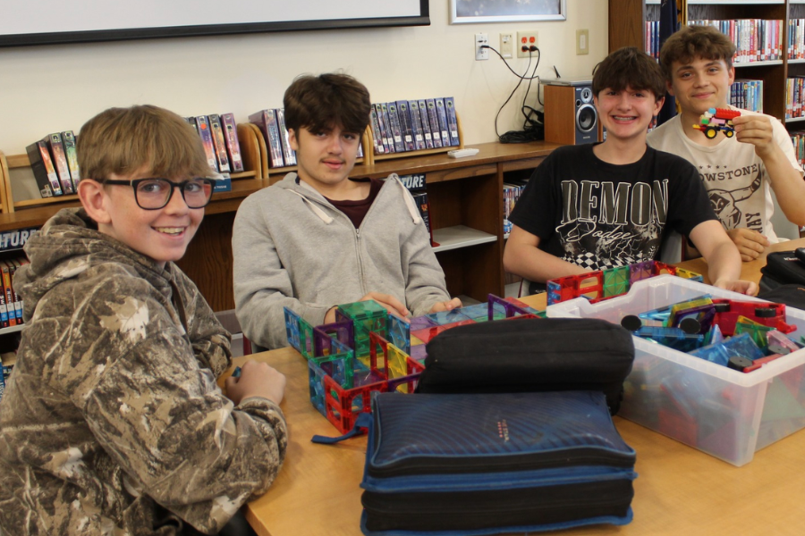 Five boys sitting at a table in a library. One holds a Rubik's cube. Behind them, shelves filled with books.