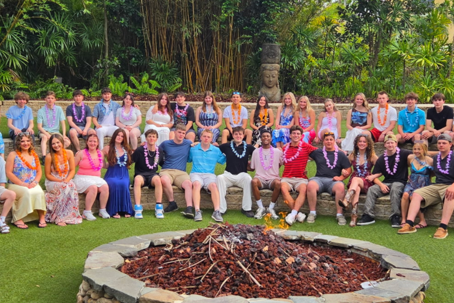 A group of people sit in tiers around a fire pit. Some wear leis. Behind them, lush greenery and a building are visible.