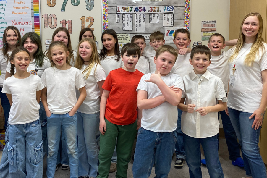 Group of students, some in white shirts, others in red, stand in a classroom. One person holds a yellow object.