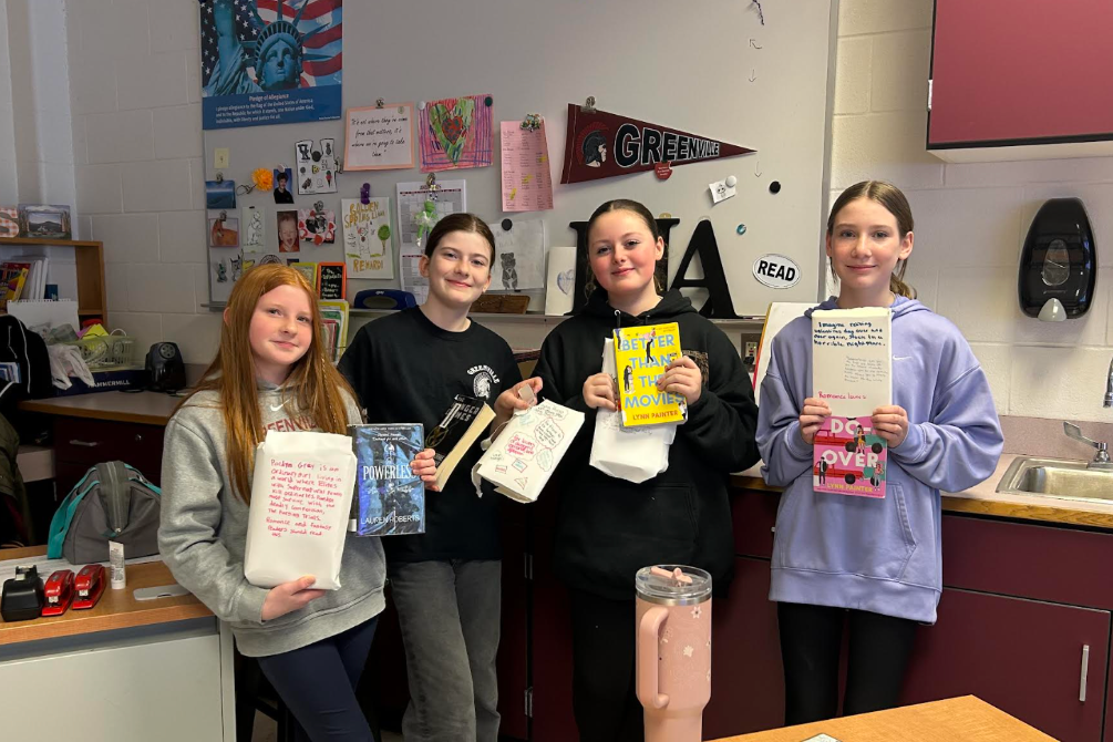 Four girls stand in a kitchen area, holding books and papers, with cabinets and a sink in the background.
