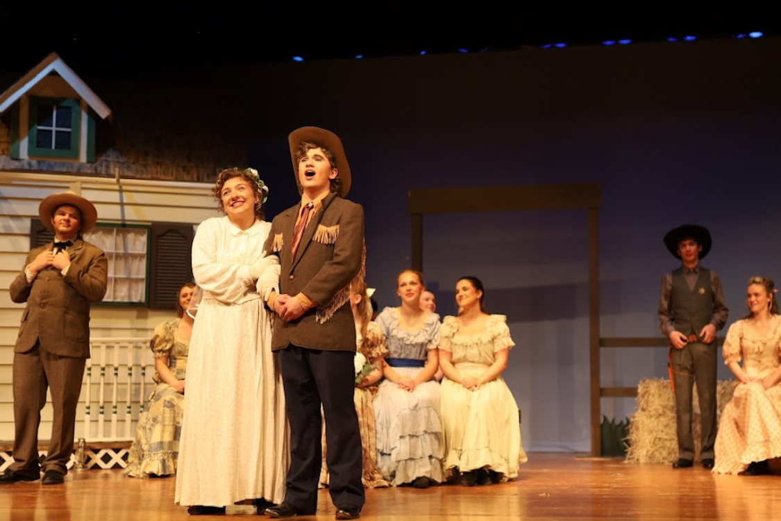 A stage with a group of people in period attire, some standing and others seated. A house and windmill are in the background.