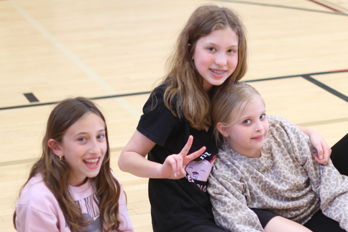 Three young girls sit together on a gym floor. One girl poses with a peace sign, while the others smile.