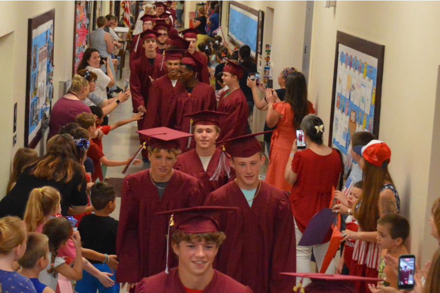 Many people in red graduation gowns, hats, and caps walking through a hallway with walls displaying artwork.