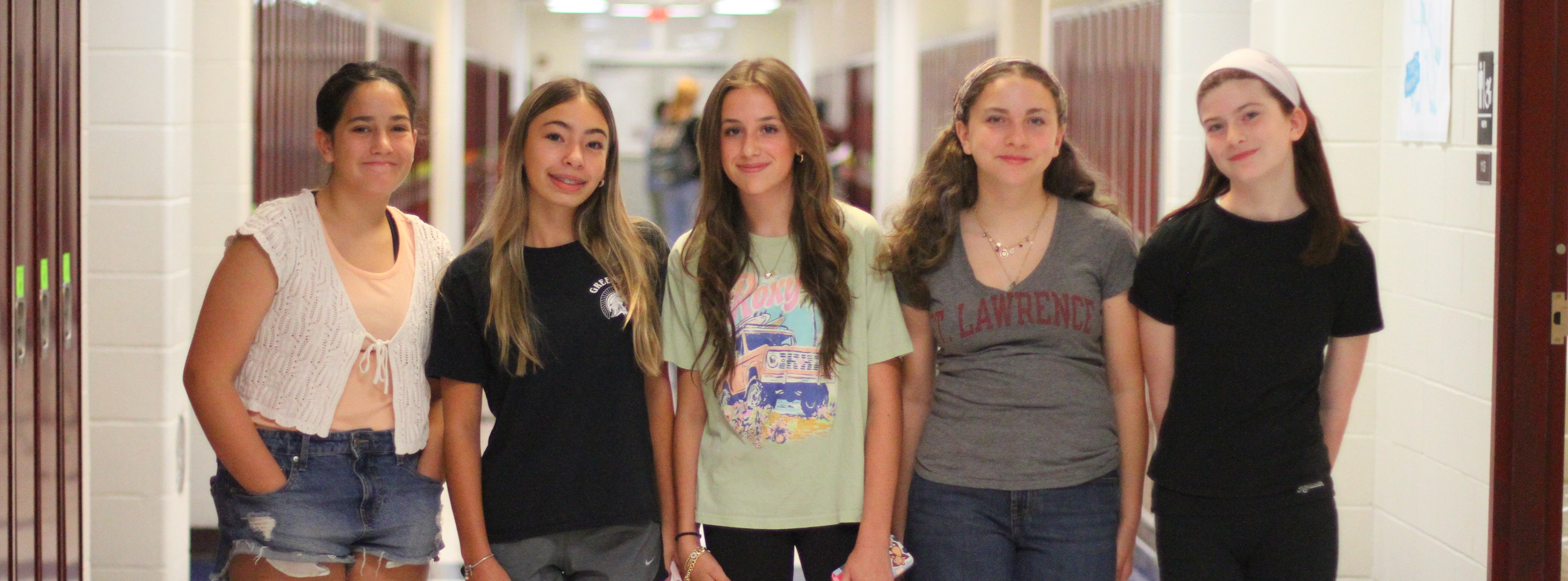 Five girls in casual wear standing in a hallway, smiling for the camera. Lockers on either side.
