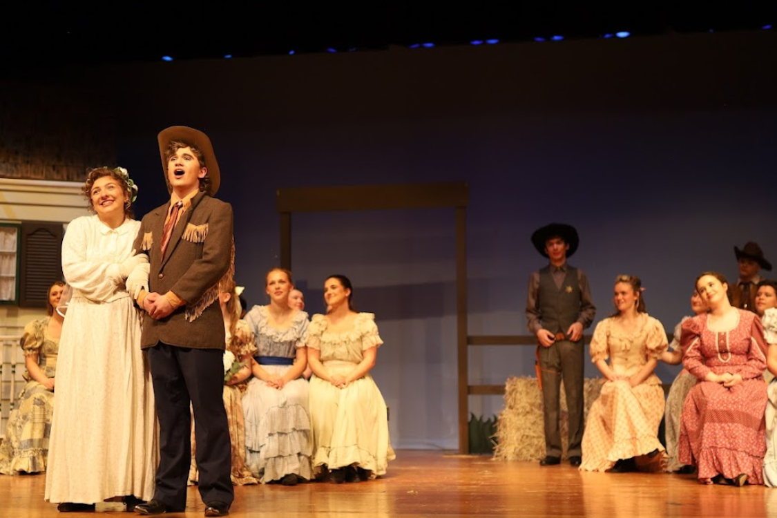 A group of people in period attire on a stage, with two central figures facing the audience.
