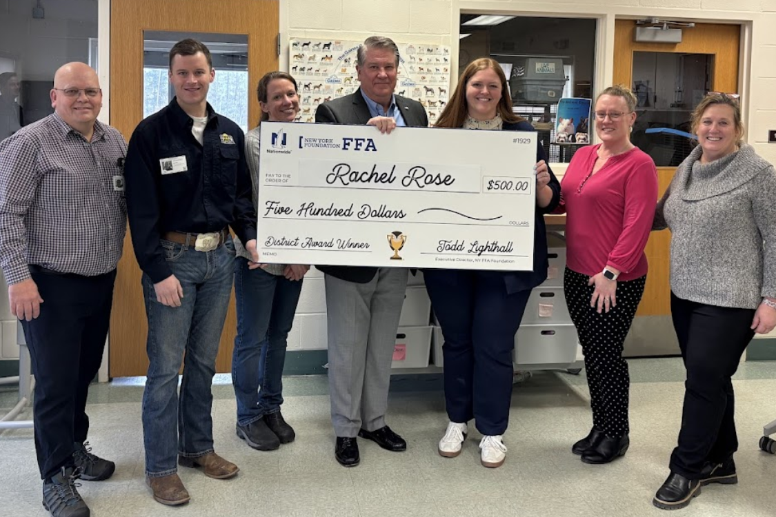 Seven people, three men and four women, stand in a room with a large check, smiles, and desks in the background.